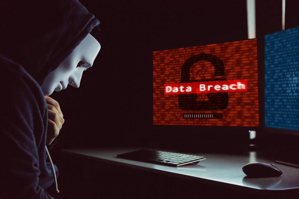 Masked hacker under hood using computer to commit data breach crime - internet computer crime concept